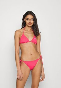 Ružičasti bikini komplet s trokutastim gornjim dijelom i donjim dijelom koji se veže sa strane. Glatki materijal s minimalnim pokrivanjem i laganim elastičnim detaljima.