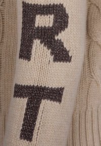 Beiger gestrickter Stoff mit großen, dunkelbraunen, metallischen Garnbuchstaben "R" und "T", die vertikal gestickt sind.