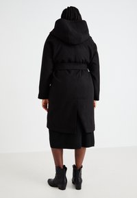 Zwarte hoodie-jack met een ceintuur in de taille, lange mouwen en een split aan de achterkant. Het materiaal lijkt dik en gestructureerd, geschikt voor koud weer.