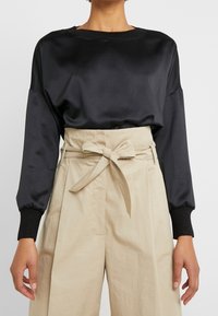 Person klädd i en svart långärmad satinblus instoppad i beiga högmidjade byxor med ett tygbälte knutet i en rosett.