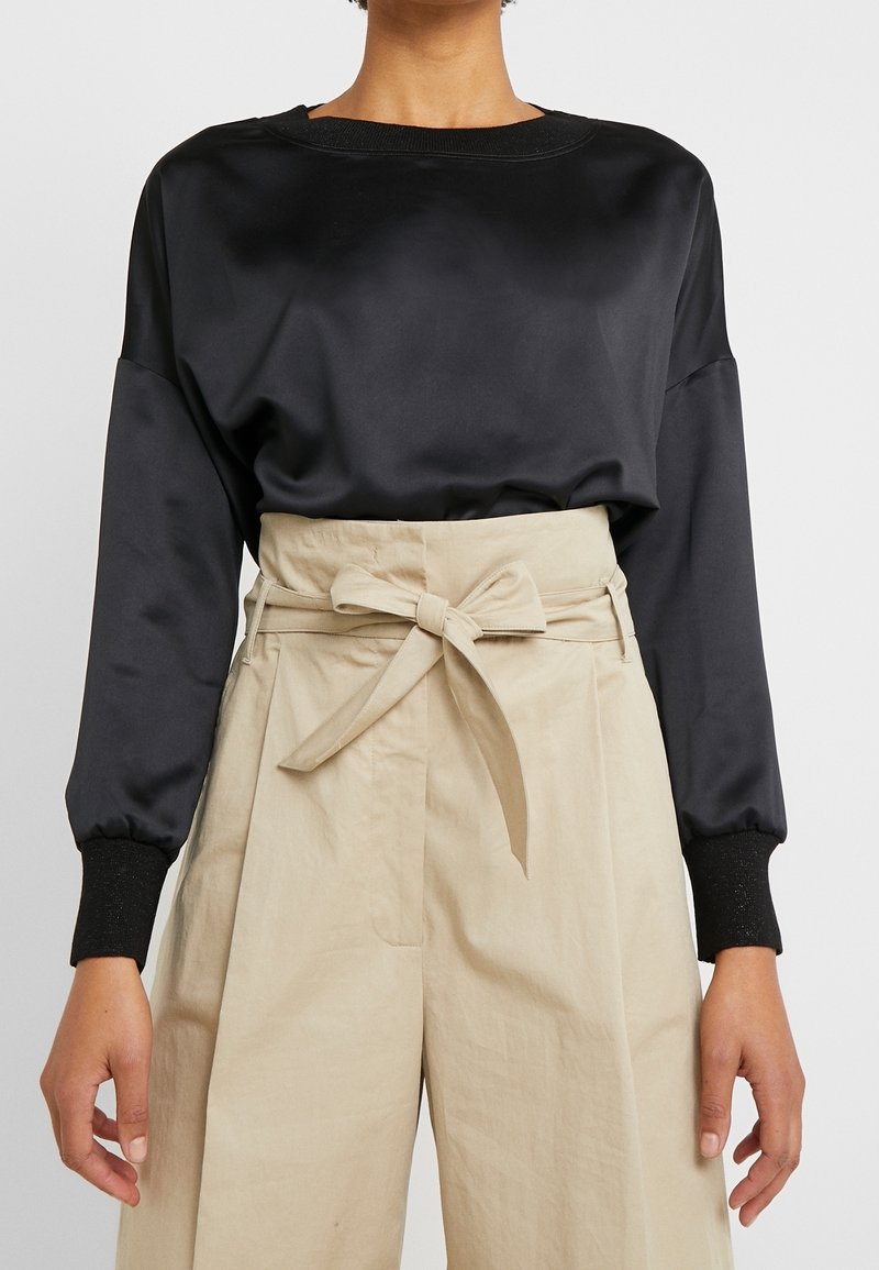 Person klädd i en svart långärmad satinblus instoppad i beiga högmidjade byxor med ett tygbälte knutet i en rosett.