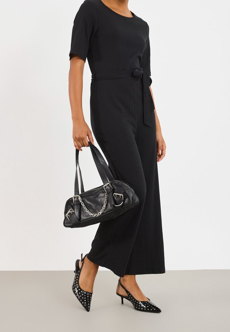 Femme en combinaison noire ceinturée tenant un sac à main en cuir noir avec détail chaîne et portant des talons slingback noirs cloutés.