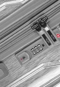 Valise rigide argentée avec double zip, serrure à combinaison et surface texturée présentant des lignes verticales.