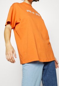 Orange bomullströja med rund halsringning och korta ärmar, med vit grafisk text på framsidan, mot en ljus bakgrund.