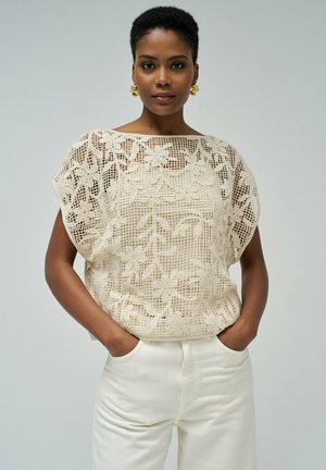 Top de encaje beige con patrón floral, de corte suelto, con un escote amplio, combinado con pantalones crema. Presenta una intrincada textura de punto y mangas cortas.