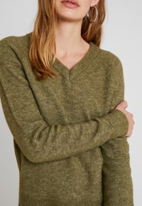 Suéter verde oliva de cuello en V, hecho de una suave tela texturizada con puños acanalados. Presenta mangas largas y un corte relajado.