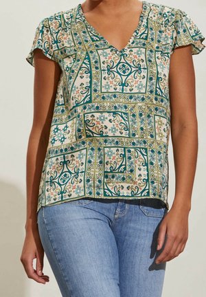 Mujer con blusa de manga corta con patrones geométricos florales en tonos azul verdoso y verde, combinada con jeans azul claro, sobre un fondo liso.