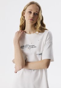 Witte katoenen t-shirt met korte mouwen, voorzien van zwarte grafische tekst en illustraties in het midden aan de voorkant. Soepele textuur, ontspannen pasvorm.