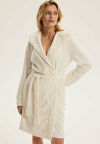 Biała frotte szlafrok z wzorem w zygzak, kapturem i paskiem w talii. Miękka tekstura, długie rękawy i średniej długości fason.