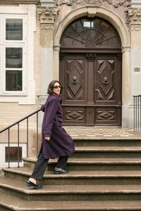 Langer lila Mantel aus strukturiertem Stoff, getragen über dunklen Hosen und glänzenden schwarzen Schuhen. Eine Treppe führt zu einer Holztür mit filigranen Details.