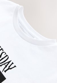 Camiseta blanca de manga corta con escote redondo y un texto en negrita negro en el frente. La tela parece suave y de textura ligera.
