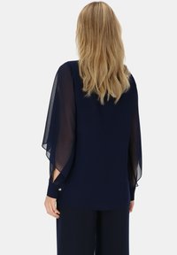 Granatowa bluzka z przezroczystymi rękawami z szyfonu, okrągłym dekoltem i subtelnymi detalami z guzikami przy mankietach. Miękka tekstura, luźny krój.
