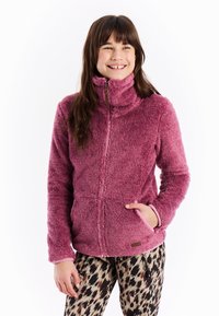 Djevojka se smiješi, nosi mekani ružičasti duksericu s patentnim zatvaračem, s rukom u džepu i tajicama s uzorkom leoparda, stojeći ispred bijele pozadine.