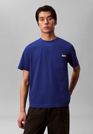 Junger Mann, der ein blaues kurzärmeliges T-Shirt mit Brusttasche und schwarze Hose trägt, stehend vor einem einfarbigen grauen Hintergrund.