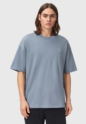 Młody mężczyzna o prostych, sięgających ramion włosach, ubrany w luźną, jasnoniebieską koszulkę z krótkim rękawem i ciemne spodnie, stoi na jednolitym tle.