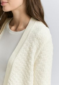 Cardigan in maglina di colore crema, con motivo texture e colletto a coste. Indossato sopra una camicia grigio chiaro, che mette in risalto il décolleté.
