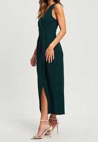 Vestido sin mangas de color verde oscuro con detalle retorcido en el frente, abertura lateral y diseño hasta la rodilla. Combinado con tacones metálicos de tiras.