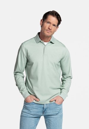 Man met kort bruin haar die een lichtgroen poloshirt met lange mouwen en lichtblauwe spijkerbroek draagt, staand met handen in de zakken.