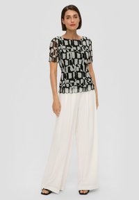 Zijdezachte zwart-witte blouse met een patroon, korte mouwen en een gegolfde zoom, gecombineerd met ruime witte broeken en zwarte sandalen.
