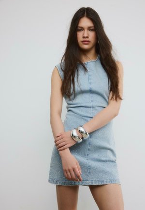 Mlada ženska z dolgimi temnimi lasmi nosi svetlo modro denim obleko brez rokavov in dve bleščeči srebrni zapestnici na levi zapestju.