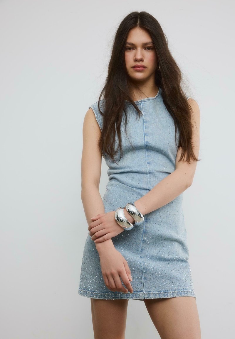 Mladá žena s dlhými tmavými vlasmi má na sebe bezrukávové svetlomodré riflové šaty a na ľavom zápästí nosí dve lesklé strieborné náramky.