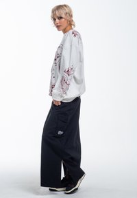 Hvid langærmet sweatshirt med rødt grafisk design på ærmerne, parret med sorte brede cargo-bukser med sidelommer. Sorte sko.