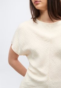 Γυναίκα που φοράει κρεμ κοντομάνικη μπλούζα με υφή και στρογγυλή λαιμόκοψη, όρθια με το ένα χέρι λυγισμένο πίσω στην πλάτη της.