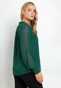 Blusa verde, texture con un motivo a pois, maniche lunghe, polsini elasticizzati e scollatura arrotondata, indossata con pantaloni neri.