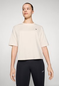 Beige cropped t-shirt met korte mouwen, ronde halslijn en een kleine driehoekige accent aan de linkerkant, gecombineerd met zwarte leggings.
