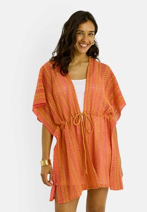 Femme souriante, portant un court couvre-chef texturé orange et rose noué à la taille sur un haut blanc, avec des bracelets dorés et des boucles d'oreilles créoles.