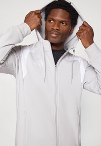 Ανοιχτό γκρι φούτερ με φερμουάρ, κατασκευασμένο από απαλό ύφασμα με κουκούλα, με λευκές λεπτομέρειες στους ώμους και κεντημένο λογότυπο στο στήθος.