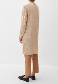 Cardigan lungo beige lavorato a maglia con vestibilità ampia, motivo testurizzato, polsini a costine e spacchi laterali, abbinato a pantaloni cropped color tan e scarpe nere.