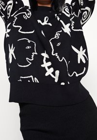 Schwarzer Pullover mit weißen abstrakten Gesichts-Mustern. Verfügt über einen runden Ausschnitt sowie gerippte Bündchen und Saum. Weicher, gestrickter Stoff.
