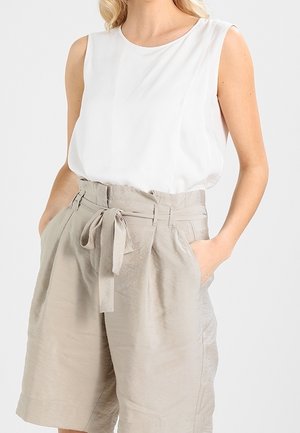 Vrouw draagt een mouwloze witte blouse, ingestopt in beige shorts die tot de knie reiken en een hoge taille hebben, met zijzakken en een stoffen riem die aan de voorkant is gestrikt.