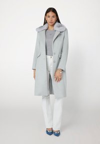 Cappotto di lana grigio chiaro con colletto in pelliccia sintetica, design aderente, bottoni frontali e tasche. Indossato sopra un top grigio e pantaloni bianchi. Scarpe blu.