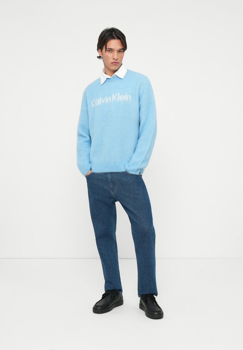 Világoskék, bolyhos kötött pulóver fehér "Calvin Klein" logóval, sötétkék farmerral és fekete sportcipővel párosítva. Egyszerű gallér részlet látható.