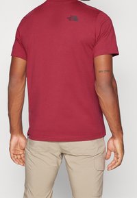 Camiseta de algodón color burdeos con mangas cortas, escote redondeado y estampado de logo negro en la parte superior de la espalda. Combinada con pantalones cargo beige.