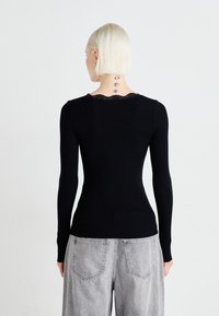 Camisola preta de mangas compridas com textura canelada, apresentando um decote em renda com bordas onduladas. Usada com jeans largos cinzentos, vista pelas costas.