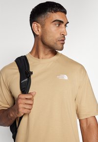 Tricou bej cu mânecă scurtă și decolteu rotund, având un logo alb pe pieptul stâng. Modelul poartă o curea de rucsac negru peste un umăr.