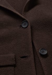 Manteau tricoté marron foncé avec deux boutons noirs visibles et une poche à rabat, montrant un tissu texturé et des boutonnières.