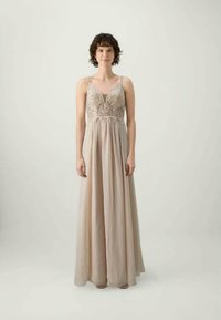 Moteris su trumpais garbanotais plaukais, vilkinti be rankovių smėlio spalvos suknelę su sudėtingais nėriniais ant liemens, stovi prieš paprastą šviesią fono.