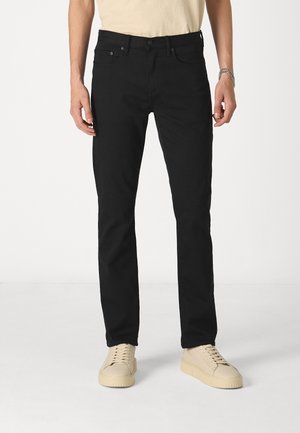 Fekete farmernadrág egyenes szabással, denim szövetből készült. Klasszikus öt zsebes dizájnnal és gombos zárással rendelkezik. Bézs cipővel viselik.