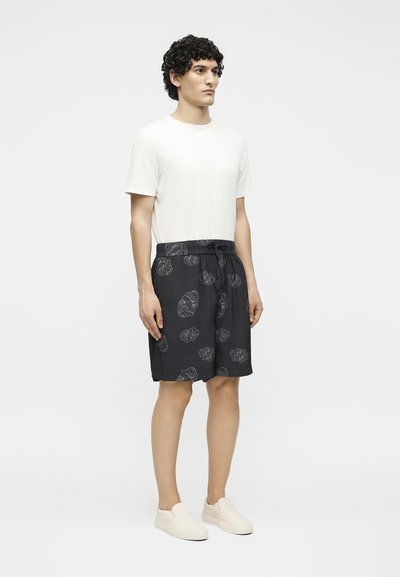 Άνδρας όρθιος, φοράει απλό λευκό t-shirt, μαύρα σορτς με μεγάλα λουλουδένια σχέδια και μπεζ παπούτσια slip-on σε λευκό φόντο.