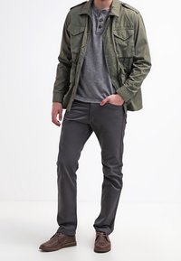 Mann steht in lässiger Kleidung mit einer olivgrünen Jacke, grauem Henley-Shirt, dunkelgrauen Hosen und braunen Lederschuhen.