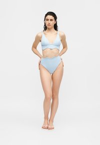 Femme en bikini bleu clair à bords festonnés et tongs blanches, debout les mains sur les hanches sur un fond blanc uni.