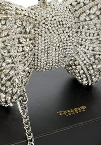 Silver rhinestone-encrusted bow-shaped handbag with chain strap resting on a black Dune London box.