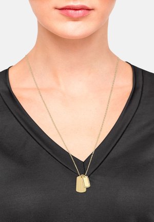 Collana con pannello per cane in oro, su una catena fine, con due pendenti dai surface testurizzati e lisci. Indossata sopra una maglia nera a V.