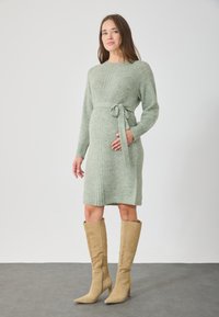 Rochie tricotată, de lungime până la genunchi, de culoare verde deschis, cu un cordon în talie și mâneci lungi, asortată cu cizme din suede bej, cu toc înalt, până la genunchi.