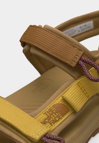 Κοντινό πλάνο κίτρινης και καφέ ρυθμιζόμενης τιράντας σανδαλιών εξωτερικού χώρου με το λογότυπο "The North Face" και μοβ λεπτομέρειες κορδονιού.