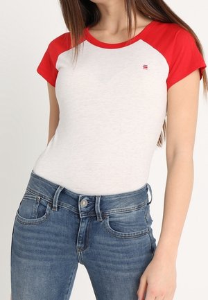 Femme portant un t-shirt blanc ajusté avec des manches et un col rouges, associé à un jean en denim bleu taille haute, debout devant un fond uni.